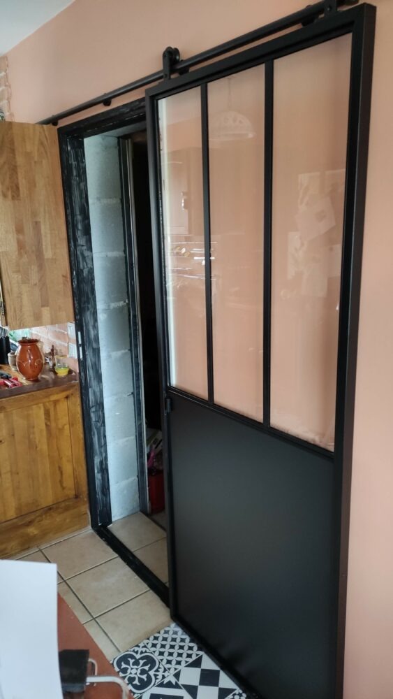 Porte coulissante noire de style atelier, partie haute vitrée en trois carreaux, en position ouverte sur son rail
